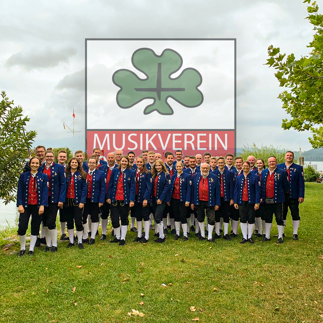 Gruppenfoto der Musiker*innen von 2019 in Tracht am Bodensee. Im Hintergrund eingeblendet das Wappen des Musikverein Salzstetten