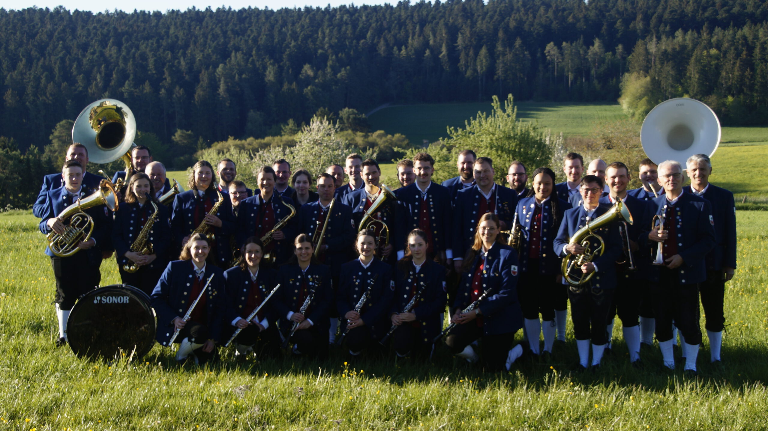 Gruppenbild des Musikverein Salzstetten. Musiker*innen in Tracht mit ihren Instrumenten in der Hand auf einer Wiese mit Wald im Hintergrund